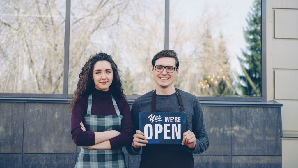 Two small business owners holding a "Yes, we're open" sign outside their shop